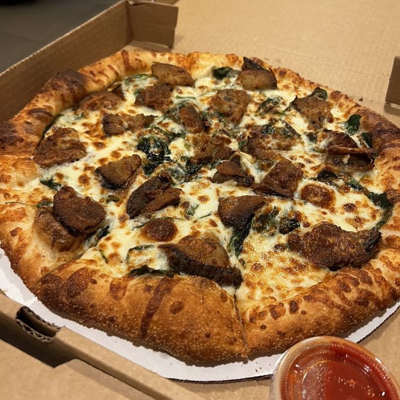 Raven Pizza's whole pizza with cheese, dark greens, and meat toppings in a cardboard box.